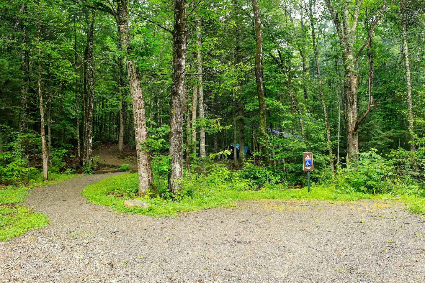 A gravel parking lot in front of a green and shaded campground. An ADA parking space is in front of the campground.